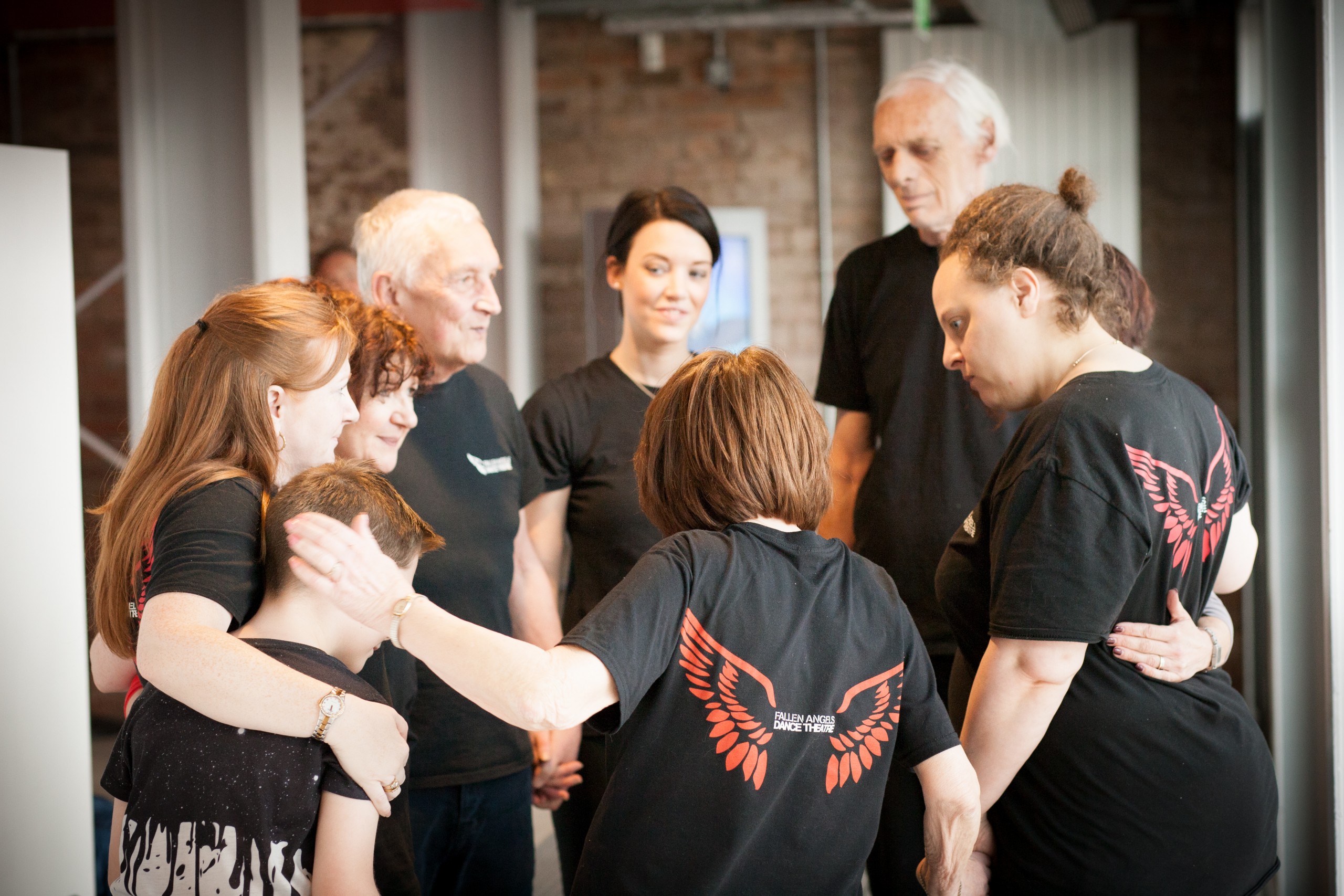 Fallen Angels dancers backstage stanfing in a circule with arms around each other. Wearing black t-shirts with red Fallen Angels wings on them.