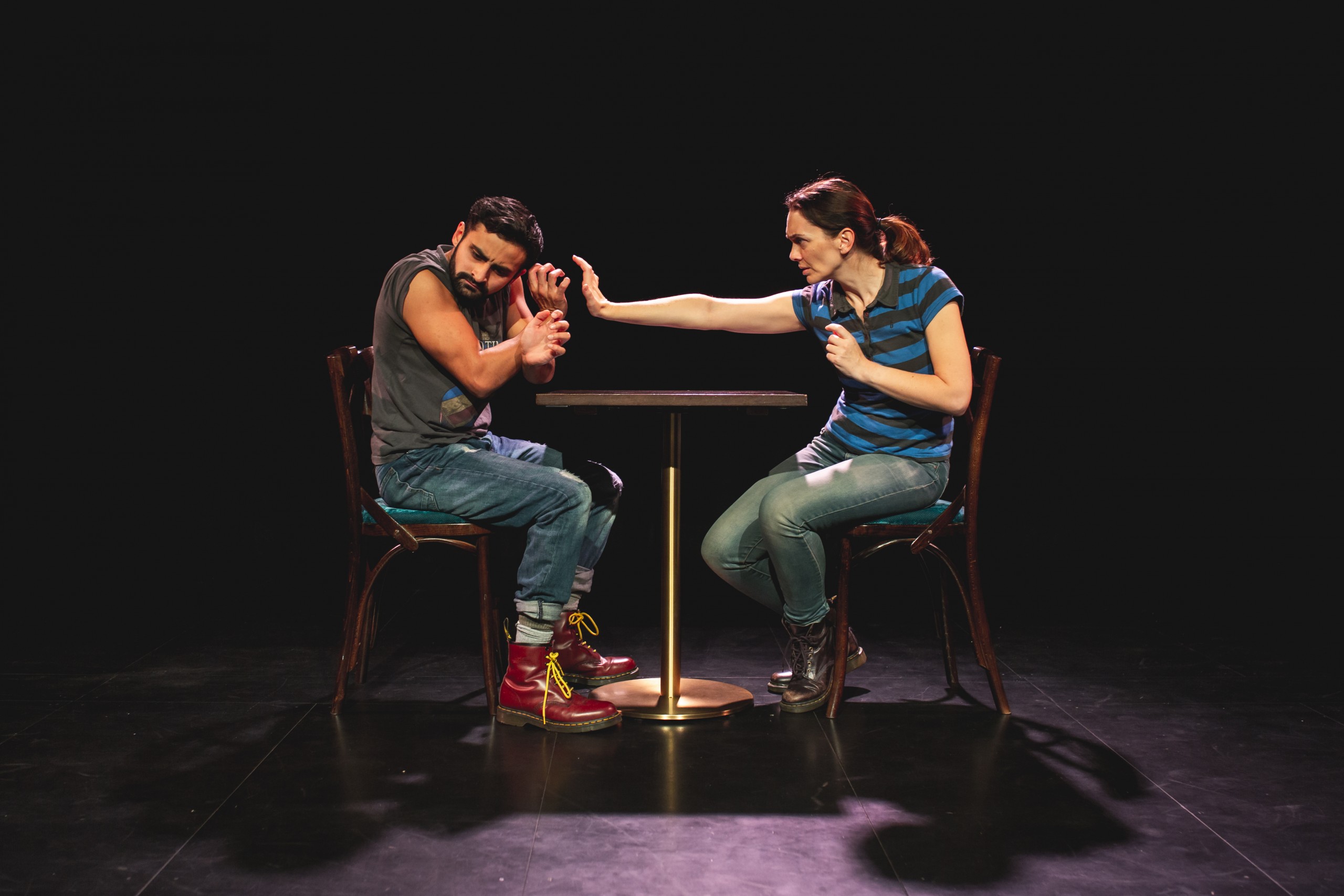 Man and Woman at a table on stage. Woman with her hand pushing towards the man's face.