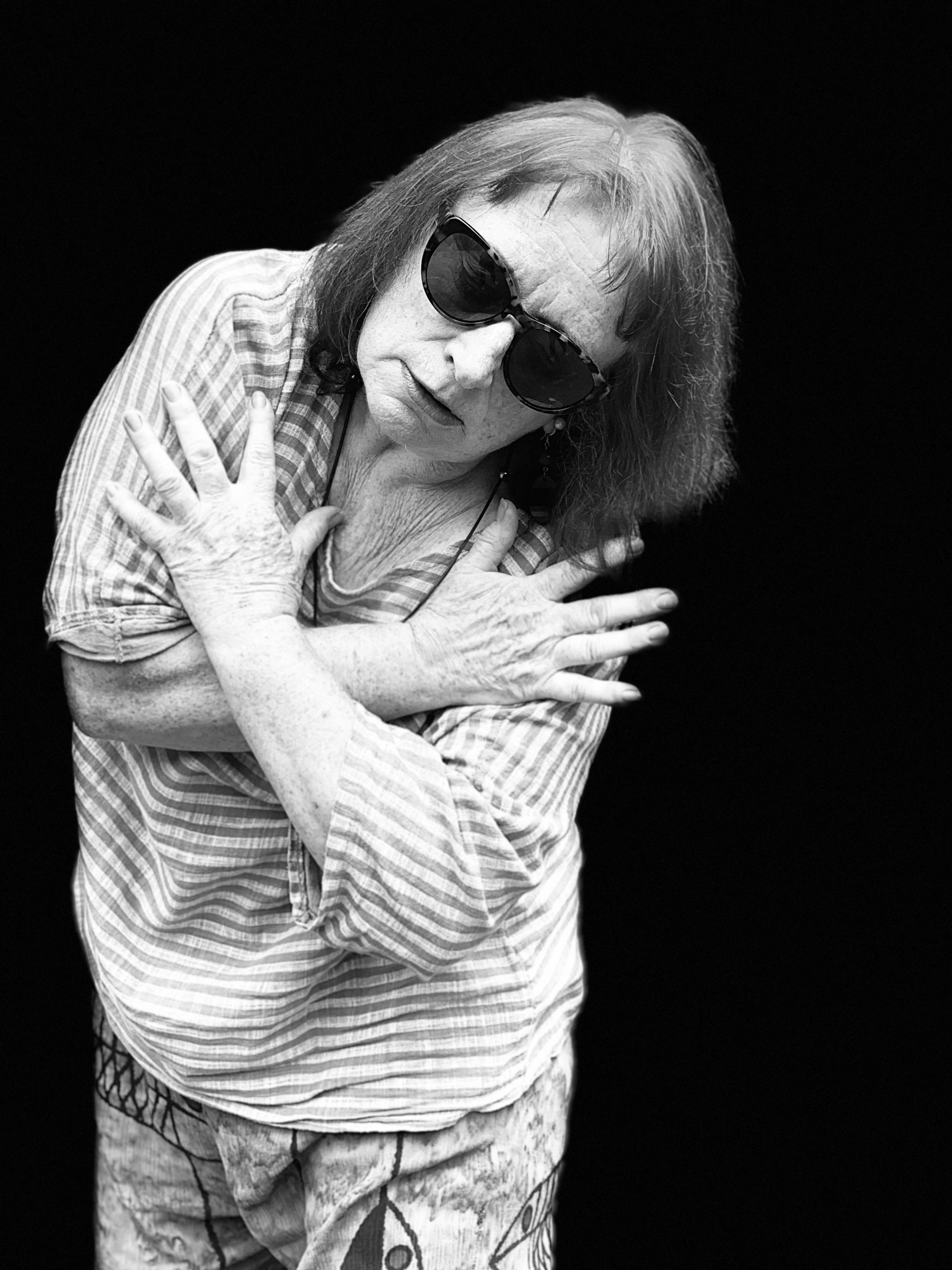 Woman wearing dark glasses, with body curved to the side and arms embraced across the body
