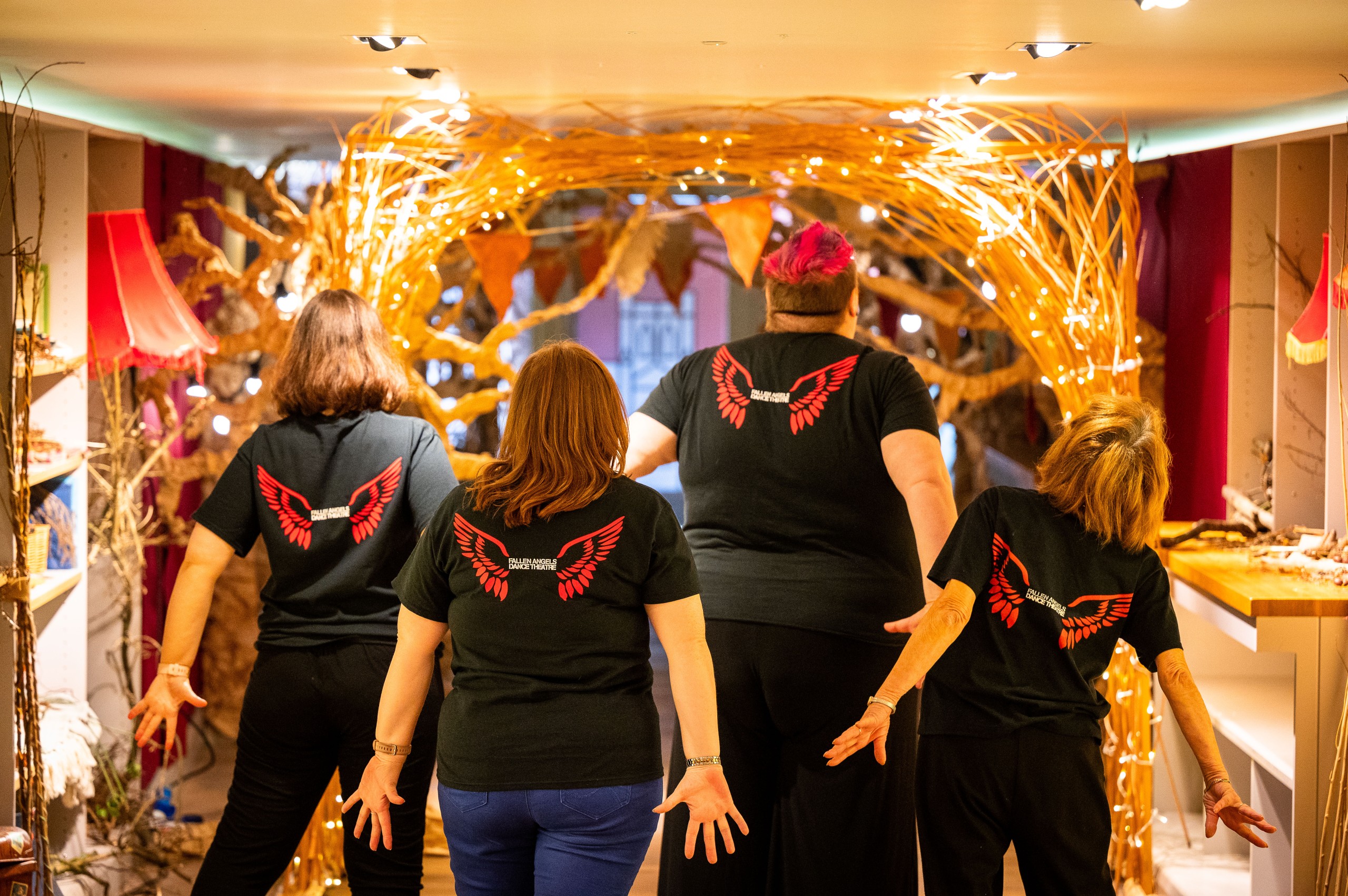 Four Fallen Angels with back to camera showing their red wings on b lack t shirts. They are surrounded by fairylights. In the Nest, Storyhouse, Chester