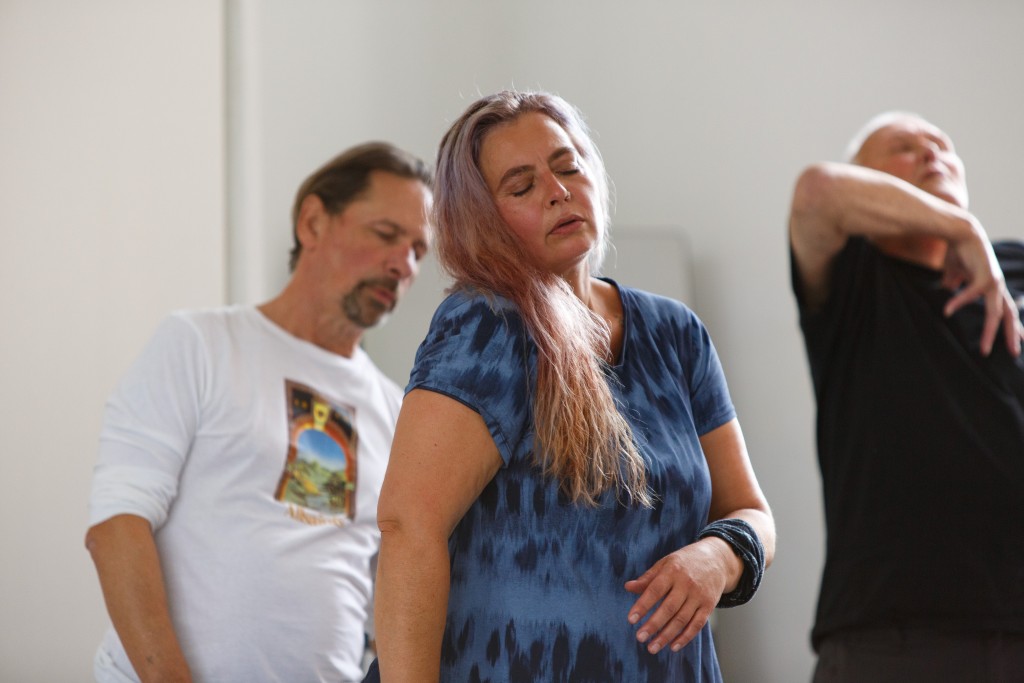 Tamara caught in motion, eyes closed as if doing a breathing exercise. One shoulder is raised with her hair draped across, one arm across her body. Two male dancers are moving behind her.